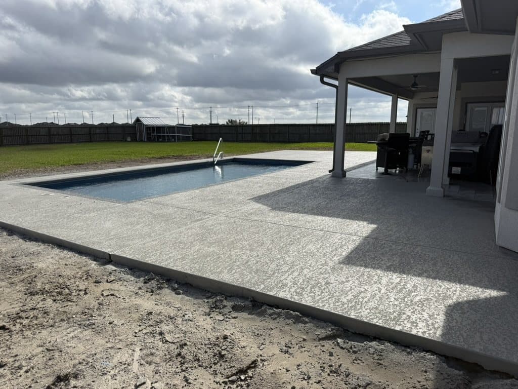 Wide light knockdown cool deck running from a covered patio out around a rectangular pool