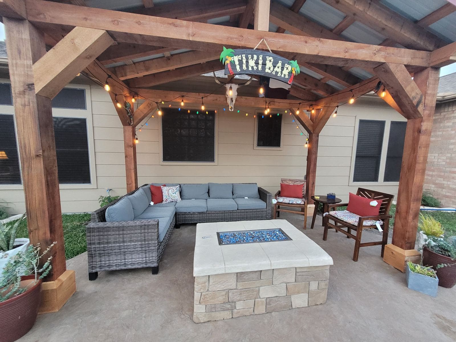 Square stone fire table under a cedar tiki bar pergola with string lights
